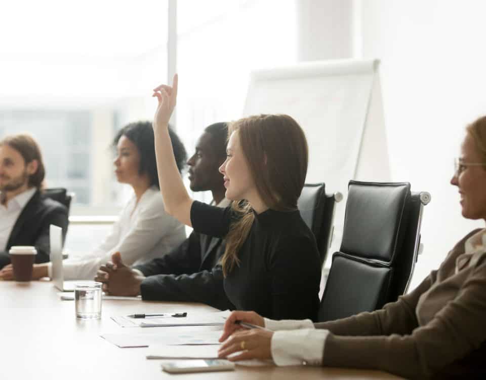Smiling curious businesswoman raising hand at group meeting